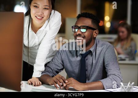 Cheerful colleagues working late in office. Young Asian woman and African American man looking at computer screen and smiling happily Stock Photo