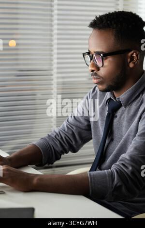 Side view of young concentrated African American office worker in glasses sitting at computer and typing on keyboard Stock Photo