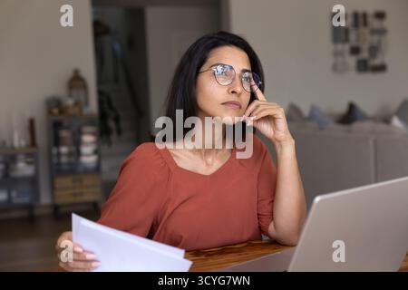 Serious pensive young Hispanic freelancer woman thinking at laptop Stock Photo