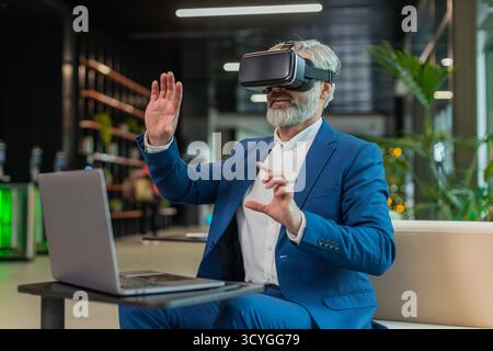 Mature businessman programmer software developer using headset app making a presentation. Watching virtual reality 3D 360 video. Bearded gray-haired guy in suit in VR goggles working at office lobby. Stock Photo