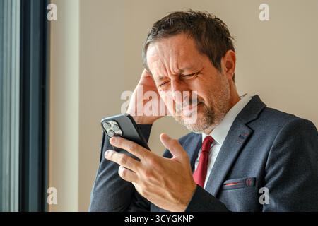 Stressed businessman reading unpleasant text message on smartphone, forgetful entrepreneur in office interior, troubled after receiving bad news at wo Stock Photo