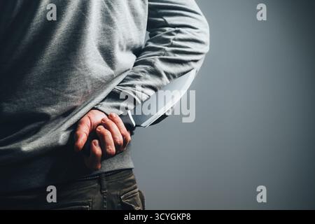 Criminal man holding large black butcher knife behind his back, symbolizing threat, danger, and hidden violent intent. Selective focus. Stock Photo