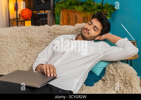 Indian young man at home, exhausted from work, falling asleep on the sofa with a laptop on his lap. Arabian guy tired lazy after long hours of work, resting on the couch, overwhelmed and drained Stock Photo