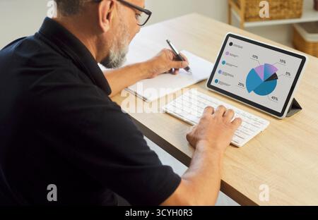 Senior man analyzing pie charts on tablet with keyboard at home office desk Stock Photo