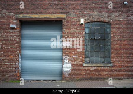 Brick factory wall with steel shuttered door and barred window in urban setting. Stock Photo