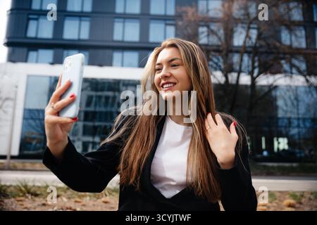 A young woman stands outdoors in a modern city environment, joyfully engaged in a video call with her smartphone, wearing professional attire and smil Stock Photo