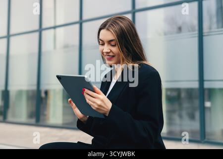A young woman in a black suit enjoys using a tablet while seated on steps next to a modern building, smiling and focused in a bright outdoor environme Stock Photo