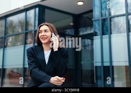 A young woman dressed in a black suit sits casually while talking on her phone outside a contemporary office building, smiling and enjoying the moment Stock Photo