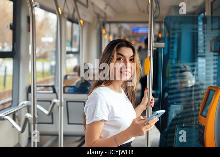 A young woman stands inside a public transport vehicle, holding a smartphone and smiling at the camera. The interior is modern and bright, typical of Stock Photo