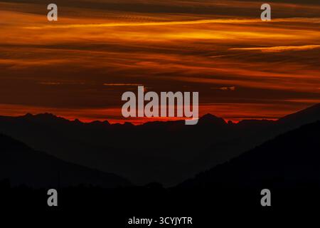 A stunning fiery sunset paints the mountain silhouettes with deep red and orange hues, creating a dramatic and peaceful alpine evening scene. Stock Photo