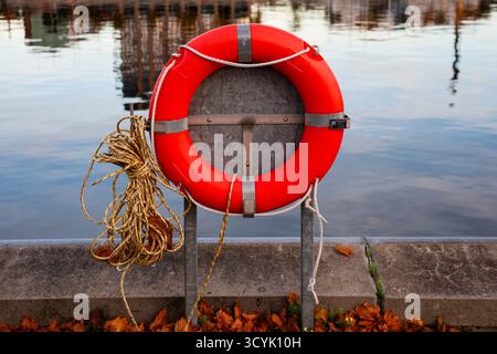 Bright orange safety lifebuoy and rope hanging on a metal stand beside the water, offering rescue and assistance Stock Photo