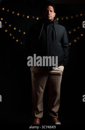 A man in a black jacket and tan pants. He is standing in front of a black background Stock Photo