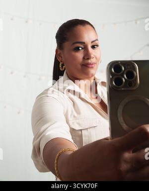 A woman is taking a selfie with her cell phone. She is wearing a white shirt and gold earrings Stock Photo