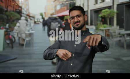 Young man with glasses smiling on a bustling urban street with people walking and cafes lining the road under a clear sky in a city setting. Stock Photo
