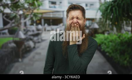Young bearded man wearing dark green shirt covers mouth with hand while standing on city street; astonishment. Stock Photo