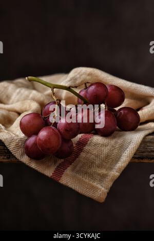 Red Grape Still Life. Closeup of  a bunch of ripe red grapes on a kitchen towel draped over the edge of a rustic wooden table with a mottled brown bac Stock Photo