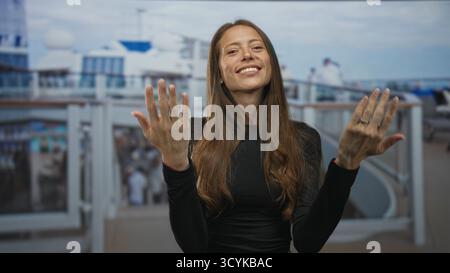 Woman beckons with both hands and open palms on building deck of cruise ship, smiling directly at camera; invitation joy. Stock Photo