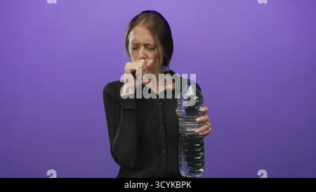 Young hispanic woman coughing into fist while holding large water bottle against a purple studio backdrop; dehydration discomfort. Stock Photo
