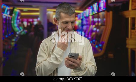 Caucasian man using smartphone in a vibrant casino, surrounded by colorful slot machines, focusing on his device indoors. Stock Photo