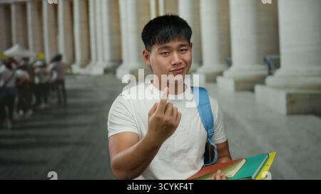 Young chinese man gestures on a city street with columns in the background, holding colorful notebooks for studying. Stock Photo