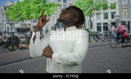 Man in white shirt pointing finger upward on street with crossed arms and serious gaze; confidence determination. Stock Photo