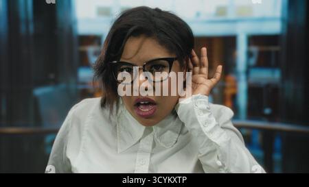 Woman wearing glasses in hotel indoors listening intently with hand on ear suggesting curiosity professional setting Stock Photo