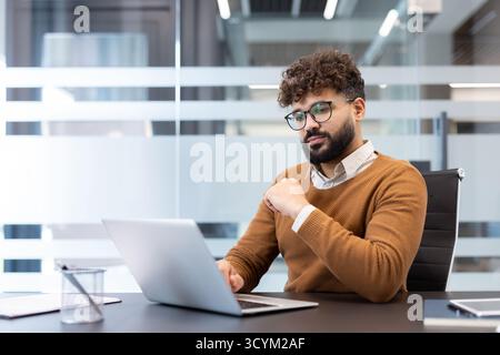 Young focused businessman working with a laptop at his desk in a blurred modern office environment, representing concentration, dedication, and professional productivity Stock Photo