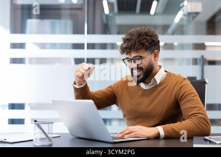 Young man with a beard and glasses raising his fist in celebration, achieving success and feeling happy while working on a laptop in a modern office environment Stock Photo