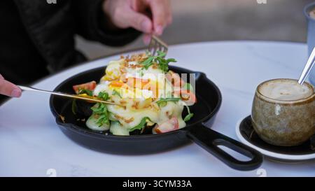Person enjoying eggs benedict on a table with cappuccino in a sunny cafeteria showcasing vibrant salad and creamy sauce. Stock Photo