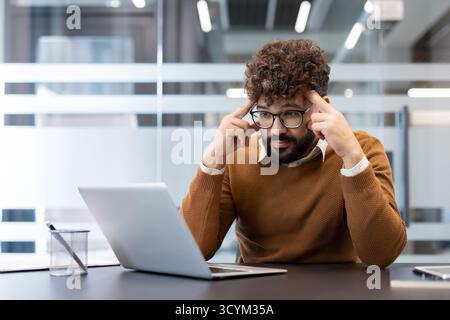 Focused young man is sitting at his office desk, displaying deep concentration and stress while thinking intently, with fingers pressed to his temples Stock Photo
