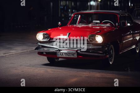 Red 1960s car on the street an night. Vintage Dodge Dart Pioneer in the dark with headlights on. Stock Photo