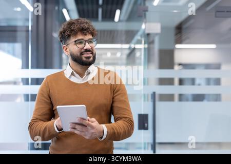 Young ethnic professional in smart-casual attire stands in a bright modern office, holding a tablet, smiling and looking away with confident, optimistic business energy Stock Photo