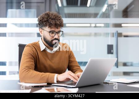 Focused young businessman with glasses and beard in a brown sweater working intently on a laptop at a modern office desk, typing and analyzing data for a project Stock Photo