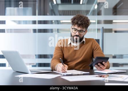 Diligent young ethnic man with glasses managing finances in a modern office, calculating budget figures and taking notes on a document with important data Stock Photo