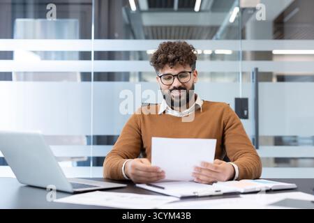 Young businessman with glasses reviewing financial reports and paperwork at a modern office desk, smiling confidently while analyzing charts, contracts and data on his laptop Stock Photo