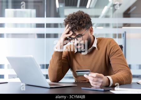 Young man feeling stressed and worried while looking at a credit card, experiencing financial difficulties and dealing with debt from online shopping on his laptop at the office Stock Photo