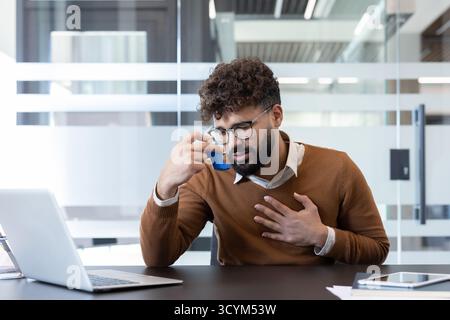 Young man experiencing an asthma attack and struggling with breathing difficulty at his workplace, using a blue inhaler while holding his chest with concern Stock Photo
