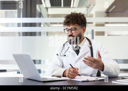 Male doctor wearing a lab coat and stethoscope is having an online video conference with a patient on a laptop, providing telehealth consultation and writing notes Stock Photo