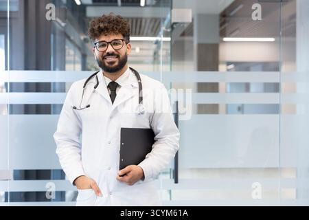 Young male doctor smiling, wearing a lab coat and stethoscope, holding a clipboard, standing in a contemporary clinic or hospital lobby, representing medical professionalism and healthcare Stock Photo