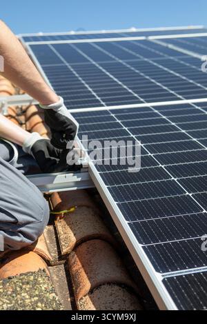 A worker in safety gear is carefully installing solar panels on a rooftop under bright blue skies. The task involves securing the panels firmly to ens Stock Photo