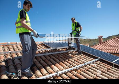 Two men are engaged in the installation of solar panels on a tiled roof under a clear blue sky. They are wearing safety vests and gloves, focusing on Stock Photo