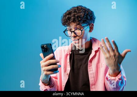 Young man in glasses uses a smartphone for video chat, showing a surprised reaction and modern online communication on a blue studio background. Stock Photo
