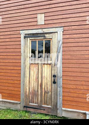 An old wooden door on the side of a rustic wood building.  The building is a dark red and the door is natural colors with three windows. Stock Photo