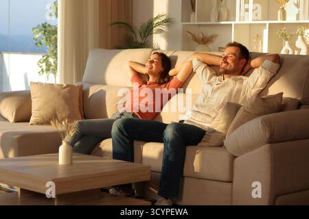 Happy couple relaxing sitting on a sofa with arms on head at sunset at home Stock Photo