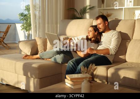 Serious couple using laptop at sunset sitting on a couch at home Stock Photo