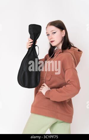 Woman musician ukulelist holding ukulele in case and looking at camera. Ukulele artist in casual clothing with brown hoodie and light green jeans Stock Photo