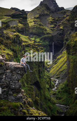 Hiker on rocky outcrop above mossy canyon and waterfall in Iceland Stock Photo