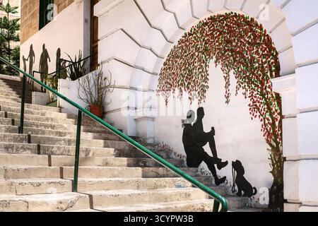 Bormla, Malta – 07.13.2025: A mural next to the staircase on a city street: black silhouettes of people and a dog sitting on the steps below. Stock Photo
