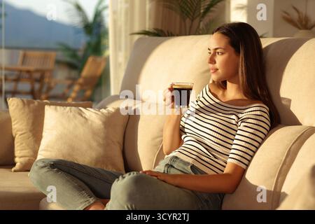 Woman relaxing drinking coffee sitting on a couch at home at sunset Stock Photo