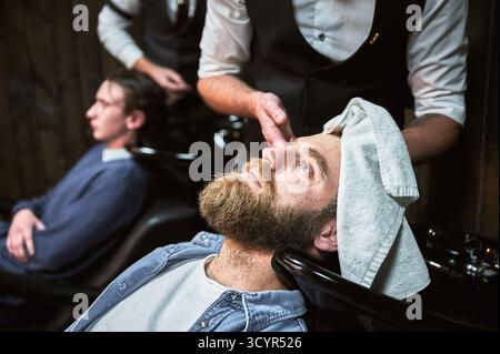 Experienced hairdresser wiping customer's hair. Close-up picture of a fancy barber drying male client's hair with white towel. Stock Photo